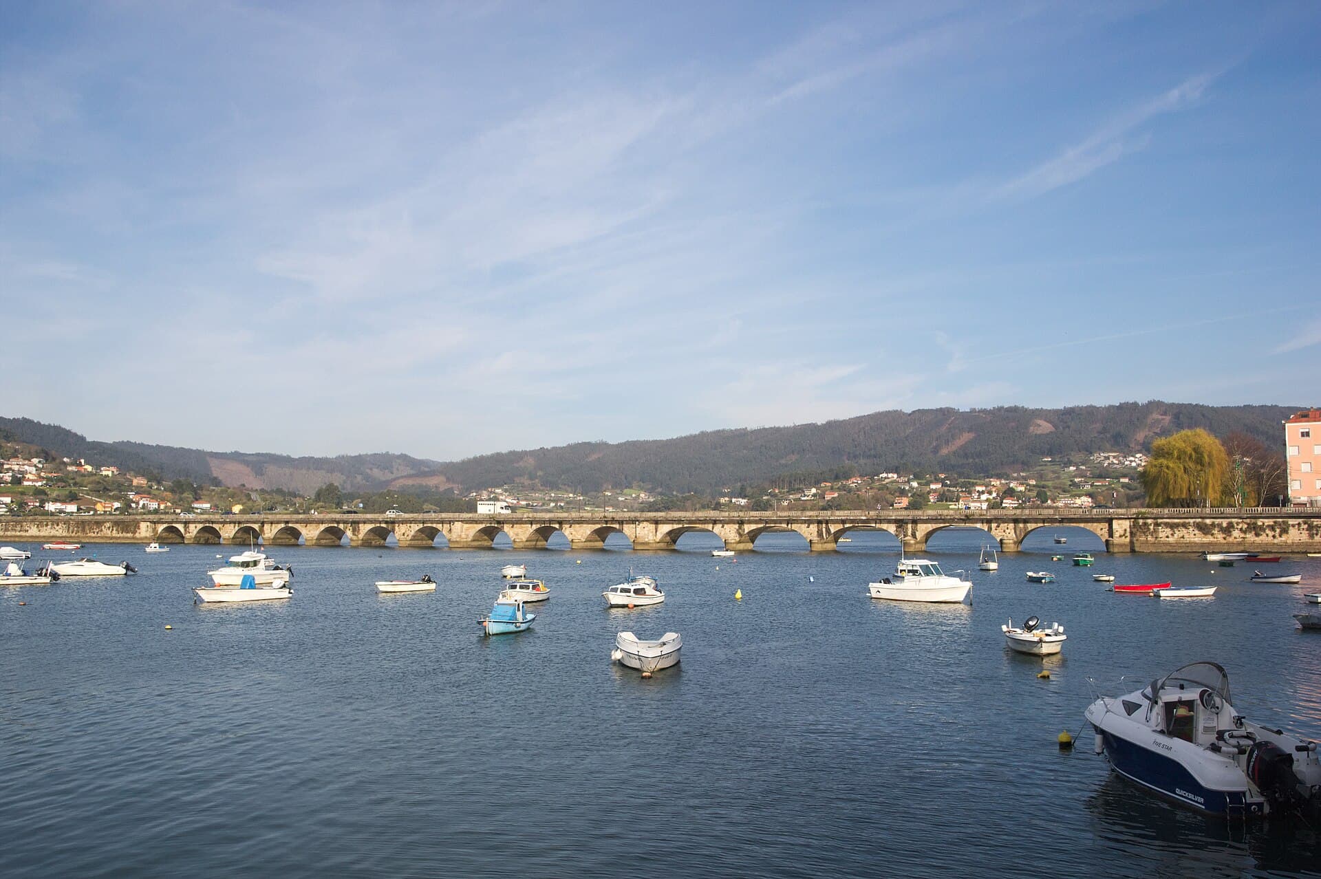 Stone bridge crossing the Eume at Pontedeume, with small boats on the water and wooded hills beyond.